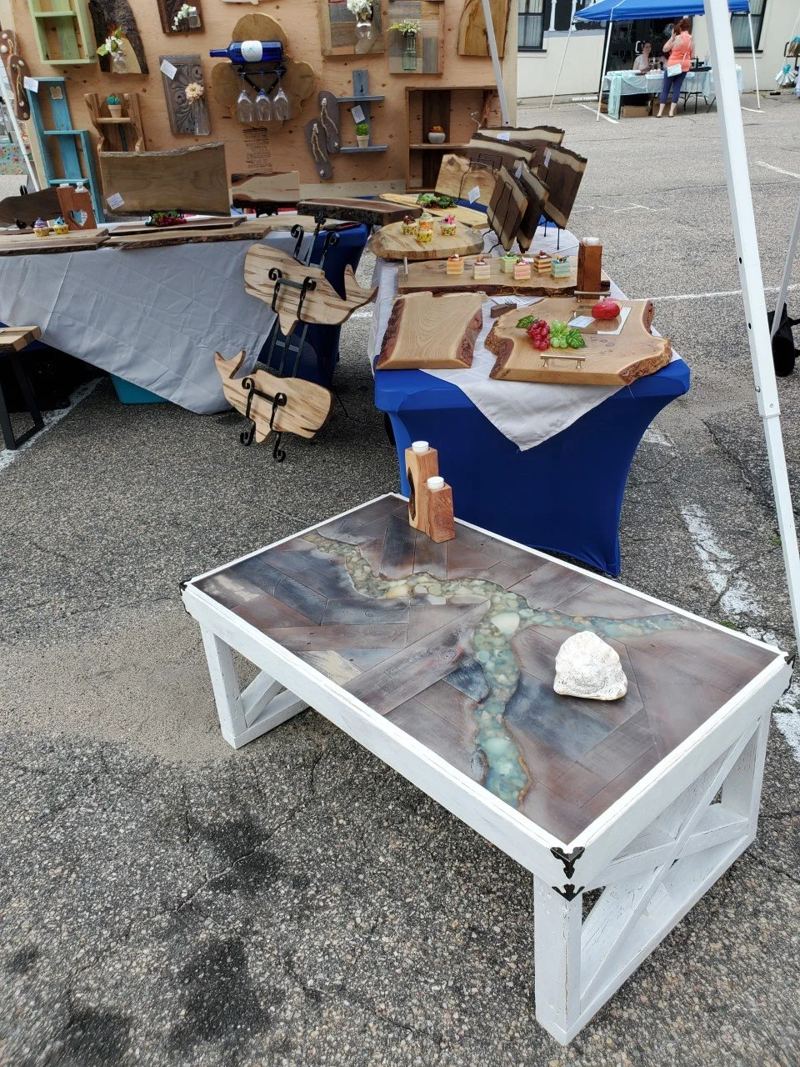 An outdoor fair booth under a white tent, with a blue river resin coffee table in front and cutting boards and signs behind.